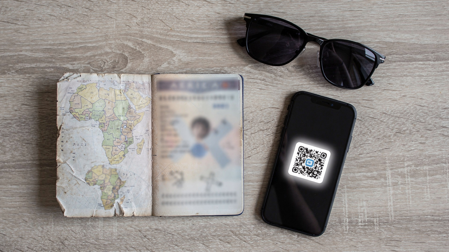 An overhead flat-lay of travel essentials: a passport, a smartphone with an eSIM QR code partially visible on the screen, a pair of sunglasses, and a small, worn map of Africa.