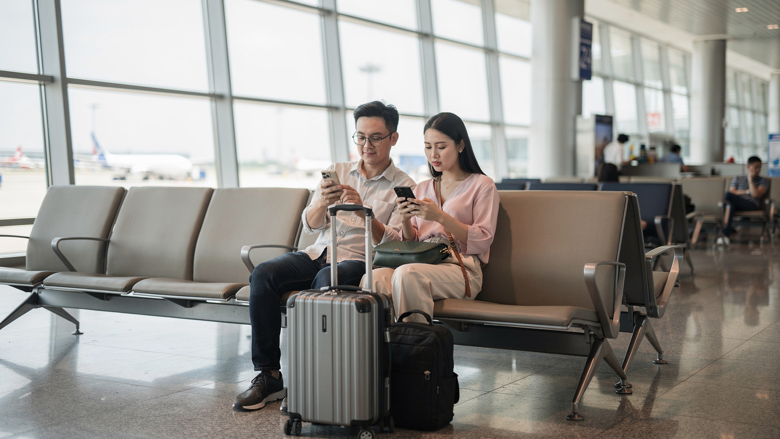 Một cặp vợ chồng trẻ Việt đang ngồi ở sảnh chờ sân bay Tân Sơn Nhất, mỗi người cầm một điện thoại thông minh. Người chồng đang xem thông tin chuyến bay, trong khi người vợ đang kiểm tra ứng dụng Cellesim để quản lý eSIM cho chuyến đi sắp tới.