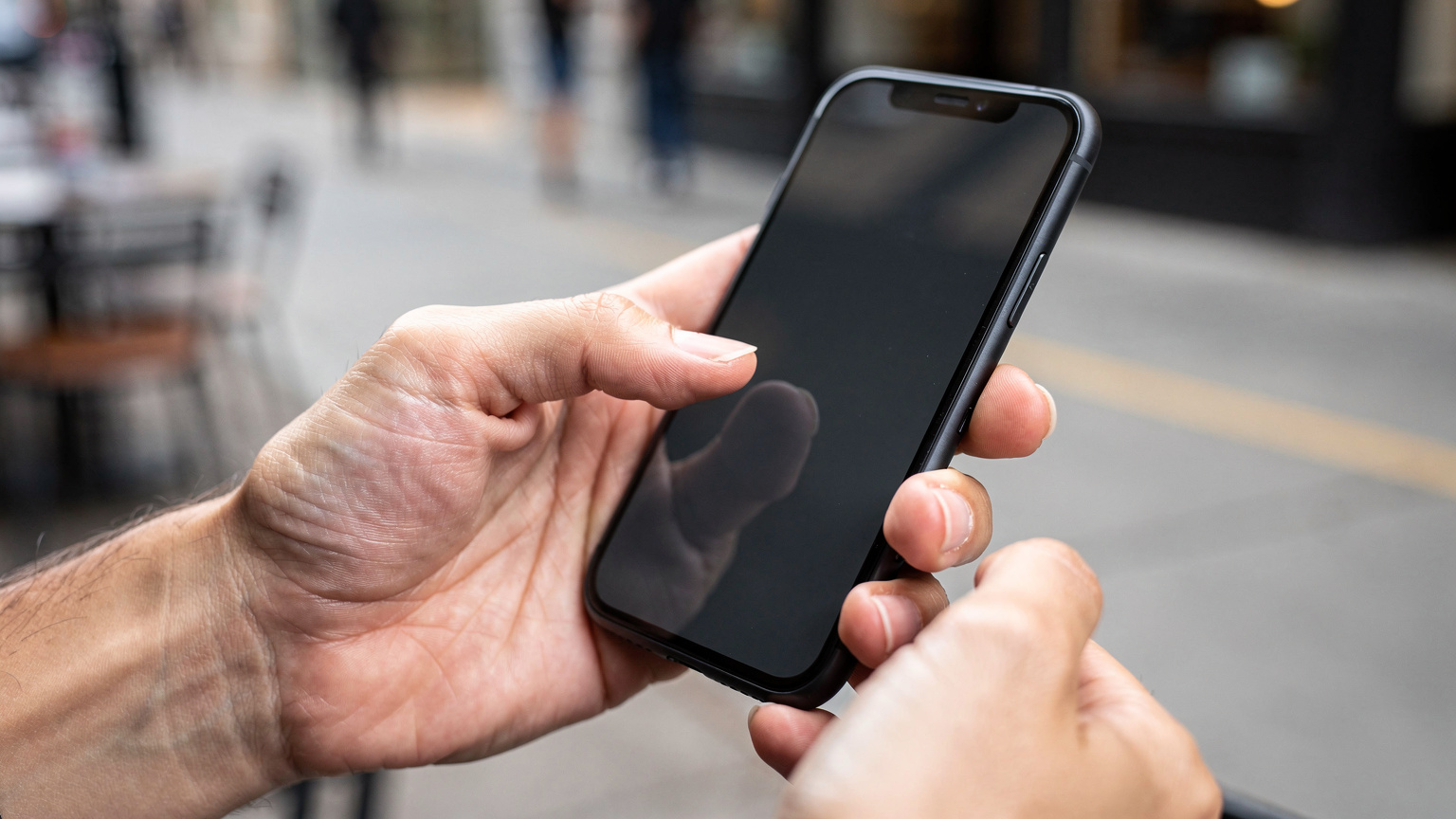 Close-up shot of a person's hands holding a modern smartphone, with one finger tapping on the screen to navigate settings, blurred city street background.