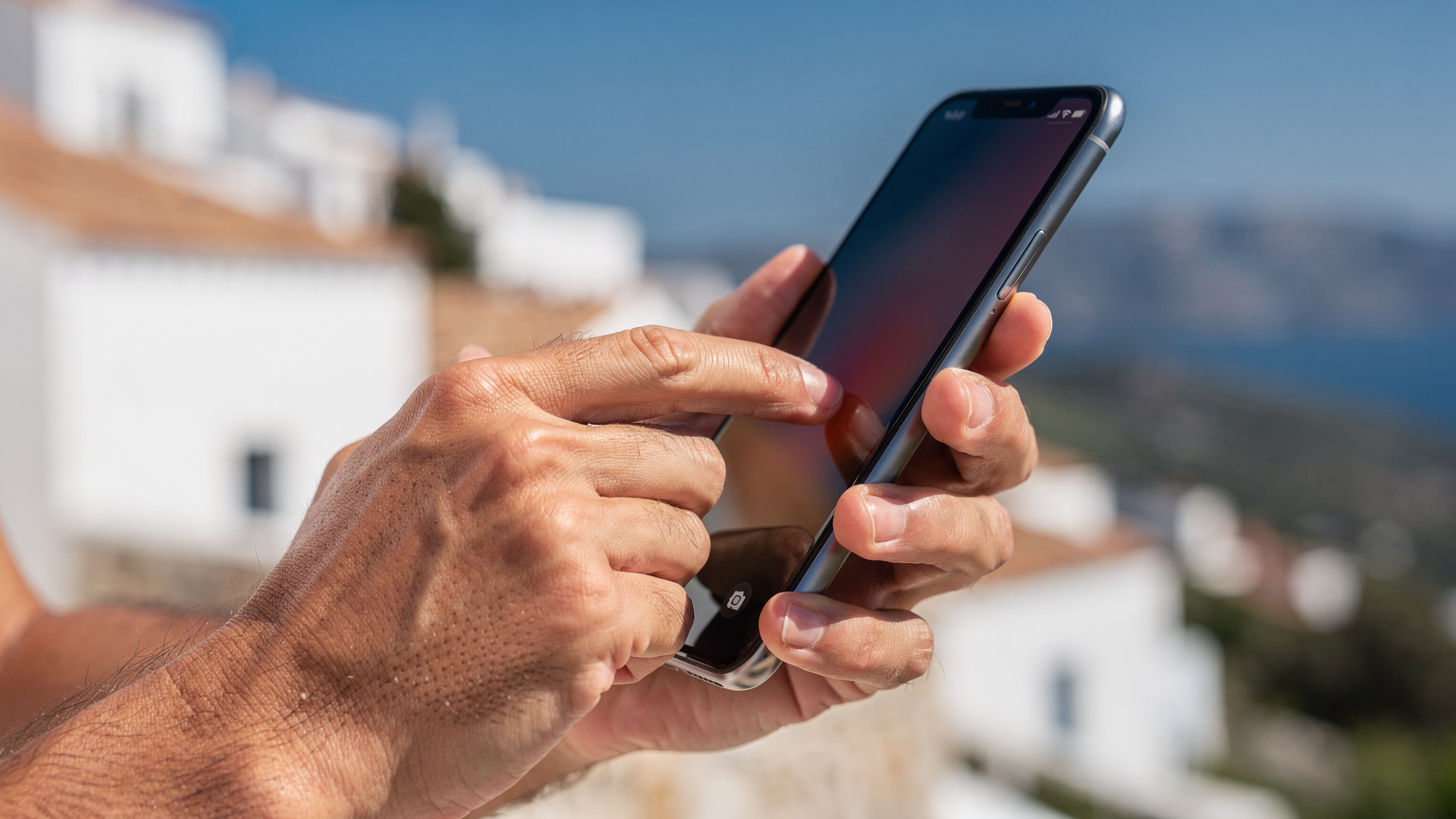 Une personne activant une eSIM sur son smartphone, gros plan sur les mains et l'écran, avec un paysage méditerranéen en arrière-plan flou.