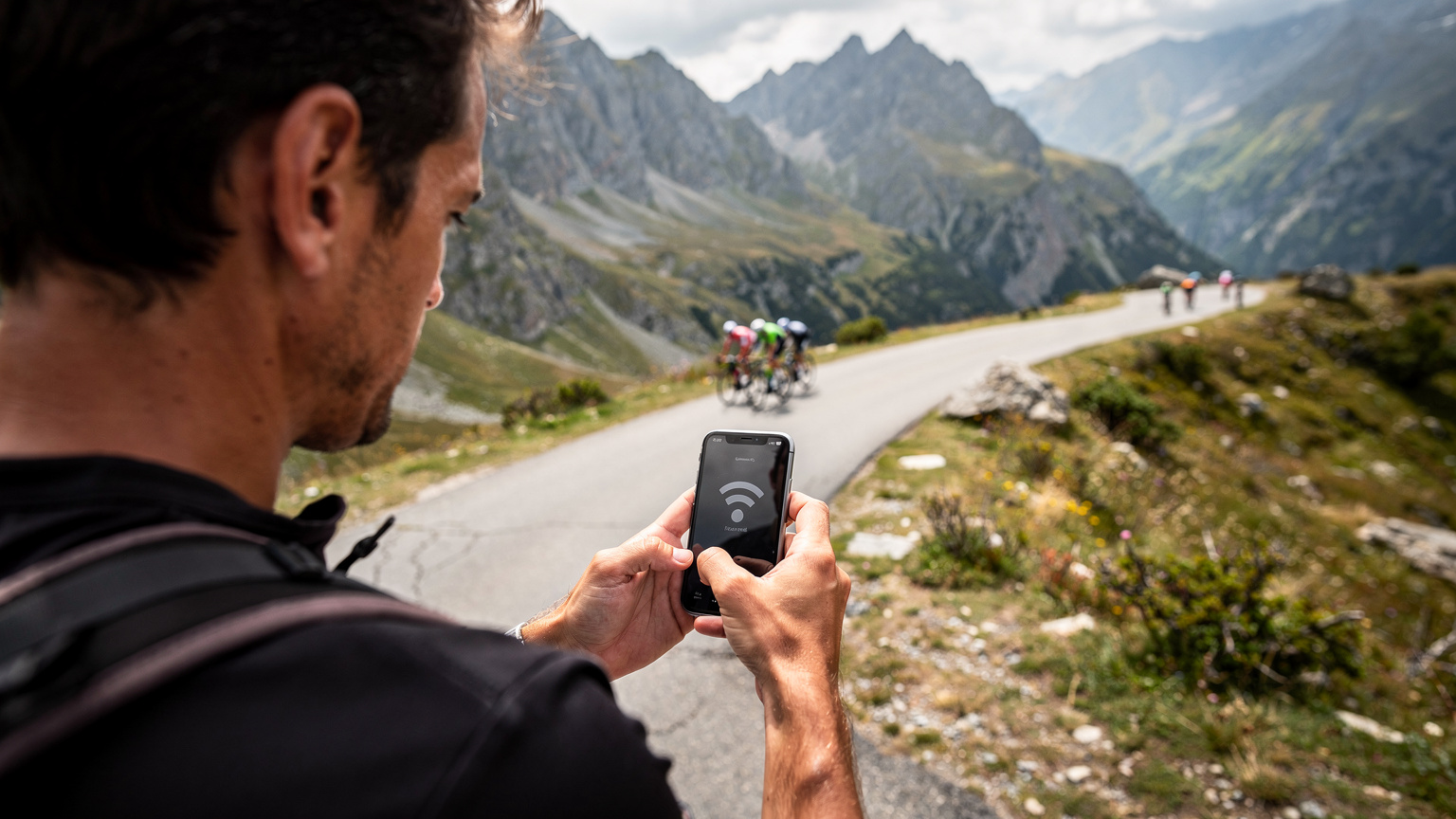 Een gefrustreerde reiziger bekijkt hun telefoon met een "Geen Service" melding, staand op een afgelegen bergweg in de Franse Pyreneeën, terwijl wielrenners in de verte passeren.