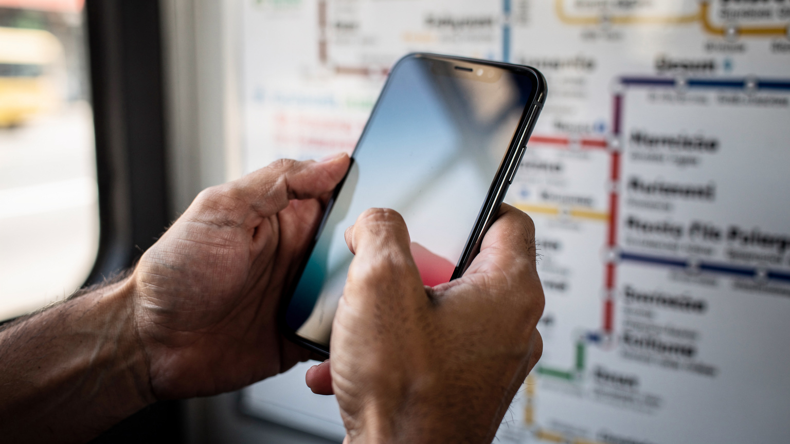 Manos de un viajero deslizando el dedo por la pantalla de su smartphone, revisando los detalles de su plan eSIM en una aplicación, con un mapa de rutas de autobús al fondo.