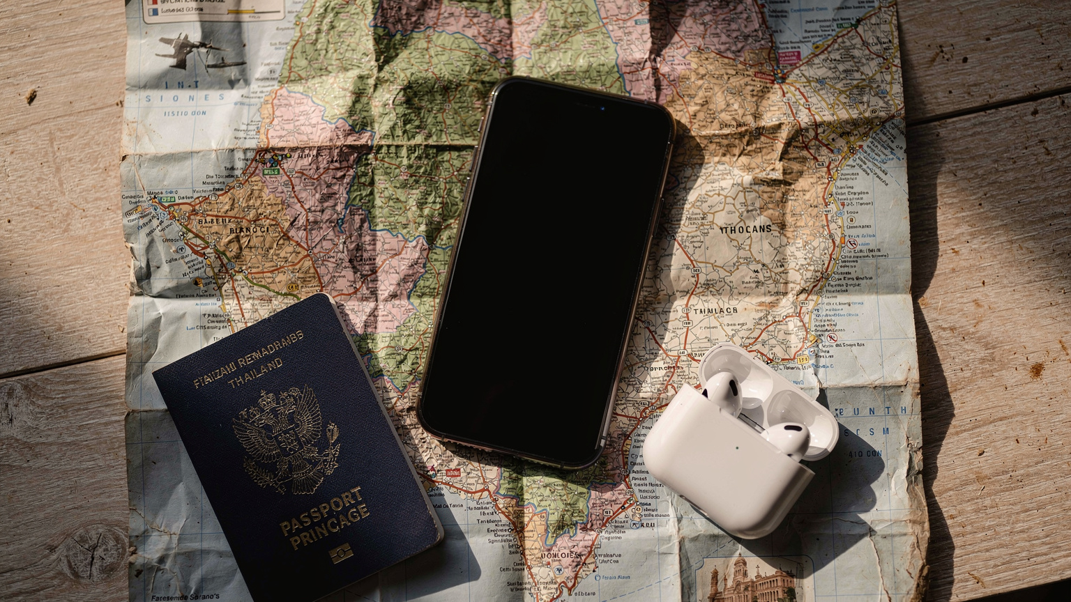 Un téléphone portable posé sur une carte froissée, avec un passeport et des écouteurs à côté, sur une table en bois clair, suggérant la préparation d'un voyage et la planification de la connectivité.
