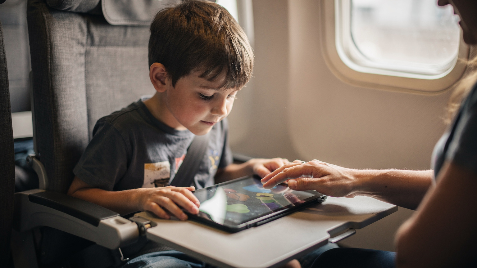 A child looking at a tablet on an airplane tray table, with a parent's hand gently guiding the screen, ensuring entertainment is ready for the flight.