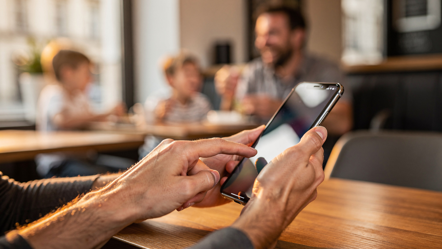 A close-up of a parent's hands expertly navigating phone settings, with a blurry background of a family enjoying a meal at a European cafe.
