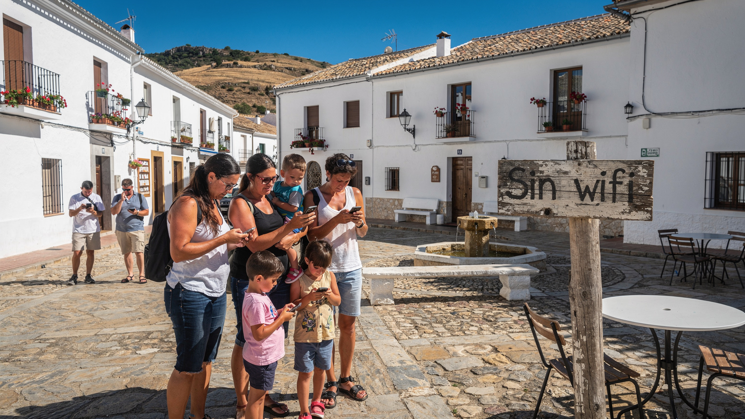 Un grupo de turistas, incluyendo una familia con dos niños, miran sus teléfonos con frustración mientras están parados frente a un pequeño letrero de 'No Wi-Fi' en la plaza central de un pueblo rural en Andalucía, España.