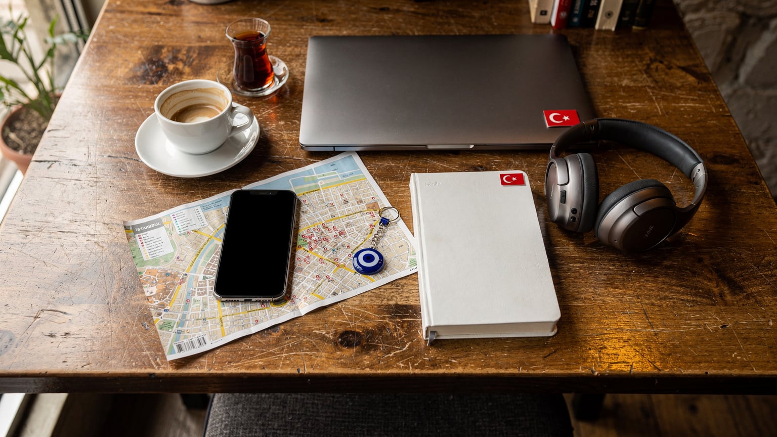 Bir dijital göçebe, bir kafede kahvesini yudumlarken, laptopu, haritaları ve not defteriyle birlikte bir masa üzerinde düzenli bir şekilde duran akıllı telefonunu gösteren bir flat-lay çekim. Yanında küçük bir Türk bayrağı detayı.