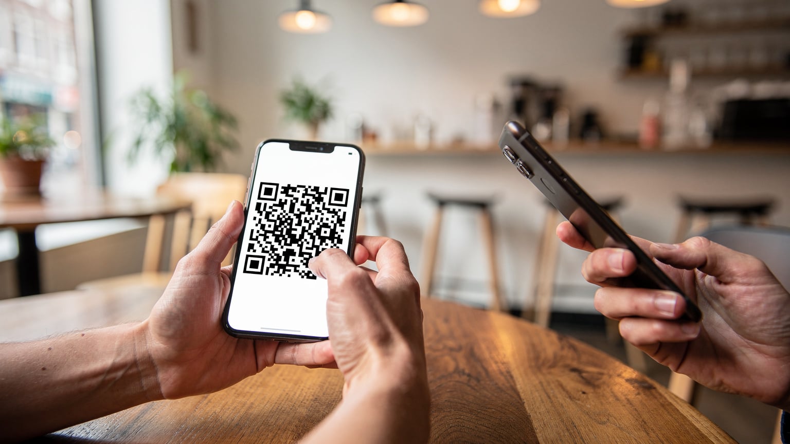A close-up of a person's hands holding a smartphone displaying a QR code, with a second phone ready to scan it, against a blurred backdrop of a bright, modern cafe interior.
