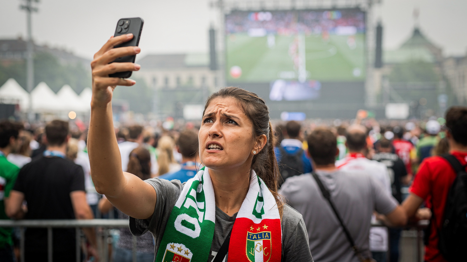 Una donna con uno smartphone in mano, visibilmente frustrata, cerca una connessione mobile in un angolo affollato di un fan park tedesco durante l'Euro 2024, con schermi giganti in sottofondo.