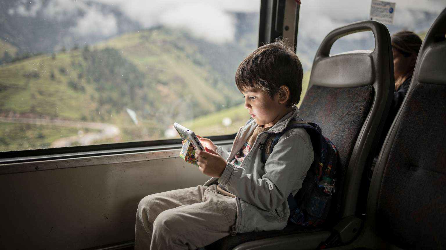 Ein Kind schaut im Rücksitz eines ecuadorianischen Busses auf ein Tablet, im Hintergrund neblige Andenlandschaft.