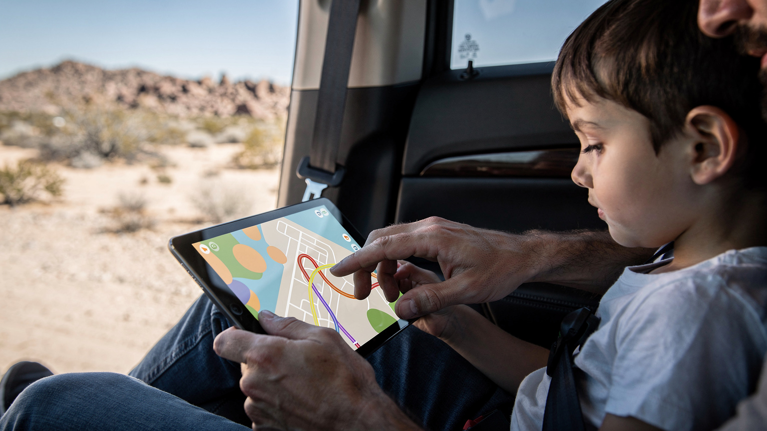 Manos de un padre y un hijo interactuando con un iPad en el asiento trasero de un coche familiar, mostrando un mapa de ruta mientras el paisaje desértico de California se ve a través de la ventana.