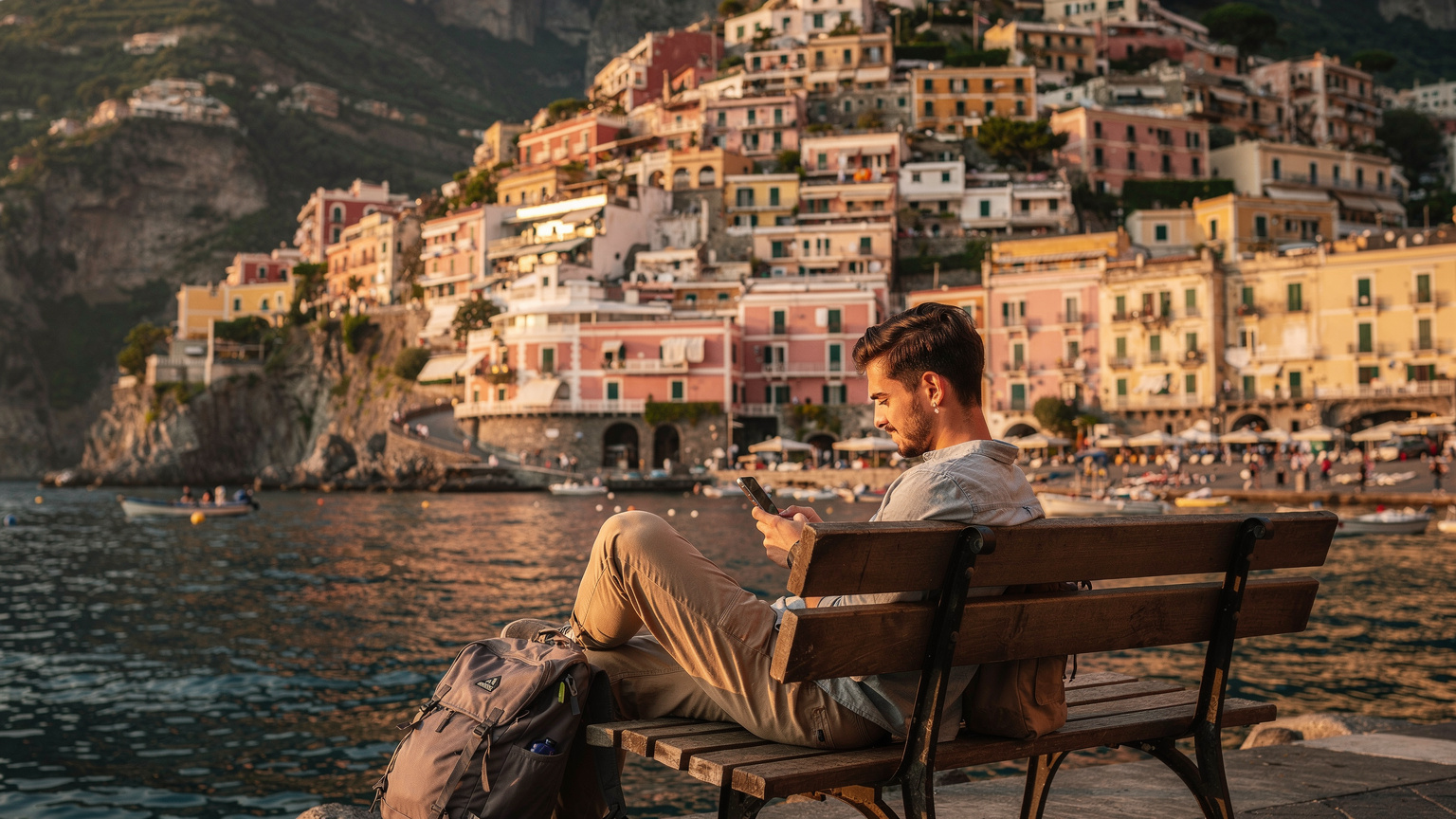 Ένας backpacker κάθεται σε ένα παγκάκι στο λιμάνι του Αμάλφι, κοιτάζοντας το τηλέφωνό του με θέα τα πολύχρωμα σπίτια της πόλης και τη Μεσόγειο.
