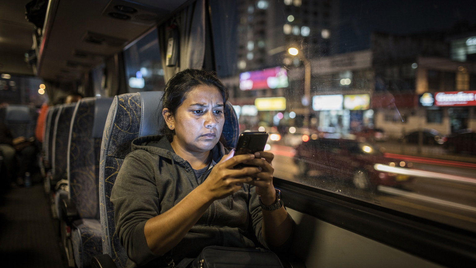 Una persona sentada en un autobús nocturno en Latinoamérica, con las luces de la ciudad a través de la ventana, mirando su teléfono con preocupación, mientras intenta conectarse a una red. Se ve cansada y algo estresada.