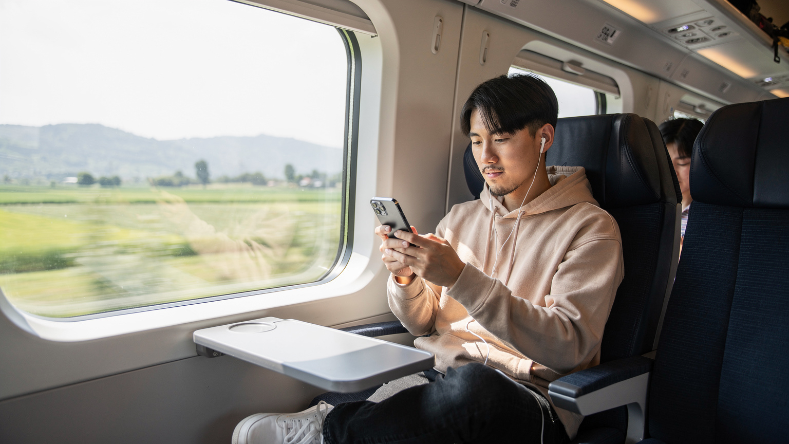 中国の高速鉄道「復興号」の車内で、窓の外に広がる田園風景を眺めながらスマートフォンで動画を視聴している乗客の横顔。