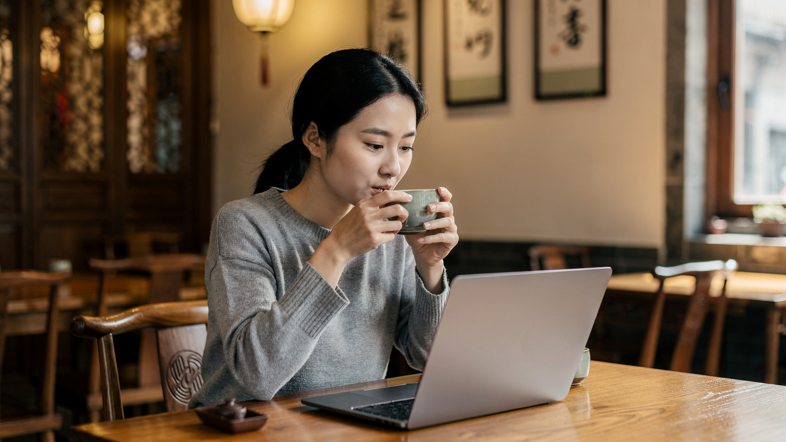 Una donna cinese, probabilmente una nomade digitale, sta bevendo tè in una tazza di ceramica e lavorando al suo laptop in un caffè tradizionale con elementi moderni a Pechino, mostrando come le persone integrano la tecnologia nella vita quotidiana.