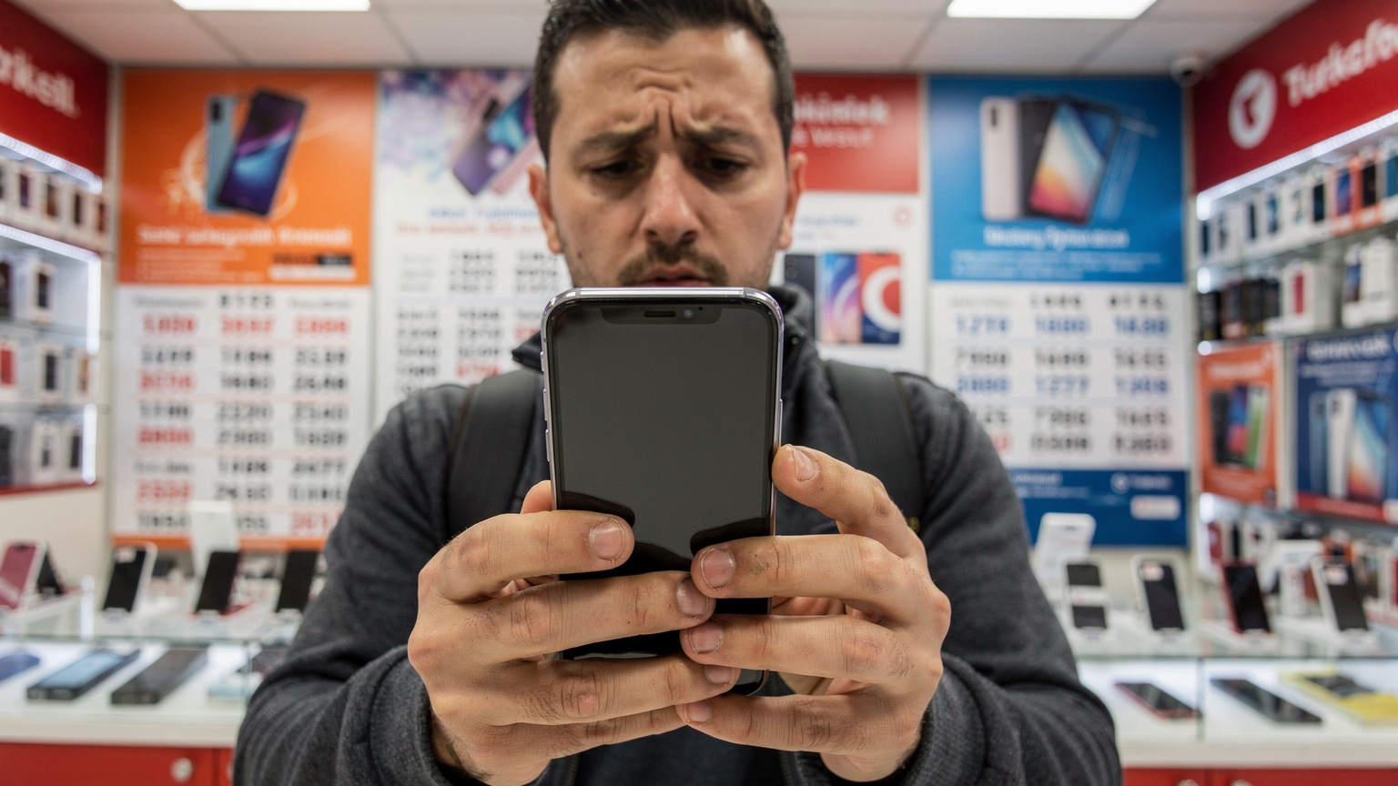 Eine Nahaufnahme der Hände eines frustrierten Reisenden, der sein Smartphone ungläubig betrachtet, während er an einem Schalter in einem türkischen Mobilfunkgeschäft steht, umgeben von Werbetafeln und verwirrenden Angeboten.