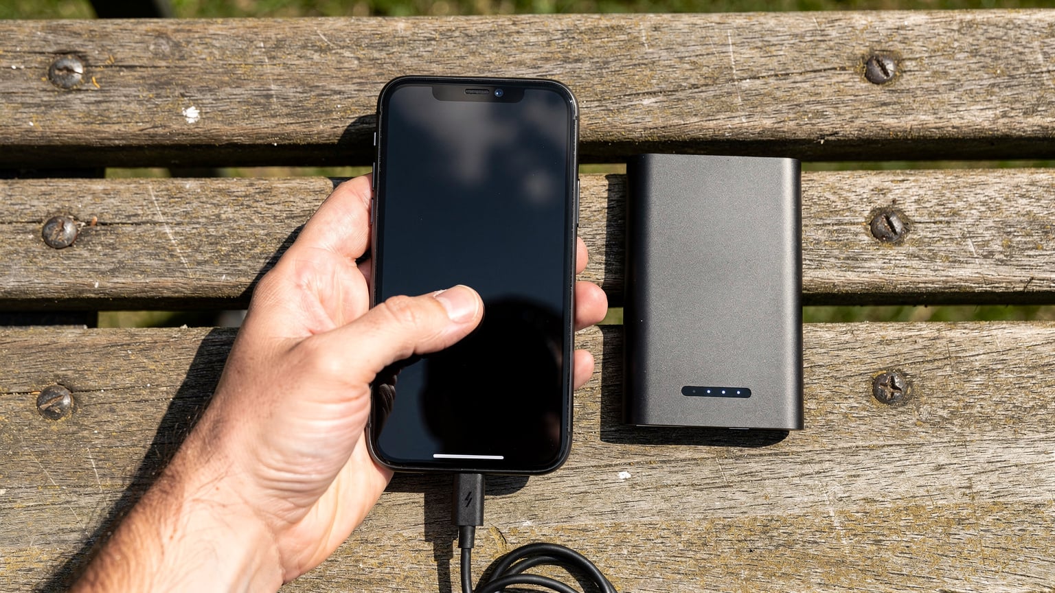 O mână de adult ține un telefon mobil, verificând nivelul bateriei sau datele, cu un power bank lângă el, pe o bancă de parc, într-o zi însorită.