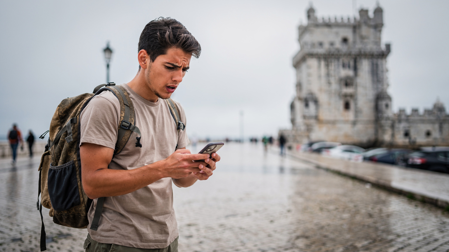 Um mochileiro confere seu celular com um semblante frustrado, enquanto a Torre de Belém, em Lisboa, aparece ao fundo em um dia nublado, simbolizando um momento de dificuldade de conexão em viagem.