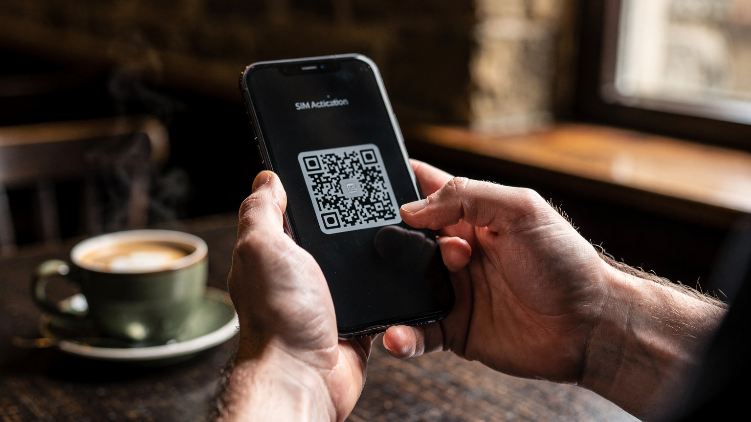 Zbliżenie na dłonie osoby trzymającej smartfon, wyświetlający ekran aktywacji eSIM z kodem QR, na tle rozmytego wnętrza szkockiej kawiarni.