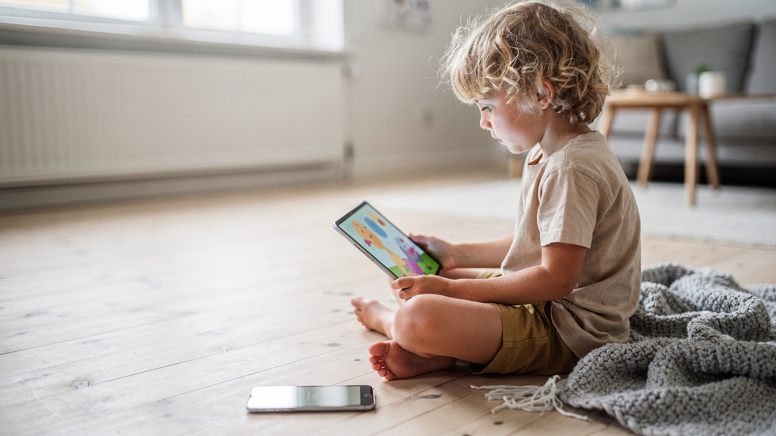 Et barn med krøllet hår holder en tablet i sommerhuset, hvor de ser en tegnefilm, med forældrenes telefon liggende ved siden af, som deler eSIM-data via hotspot.