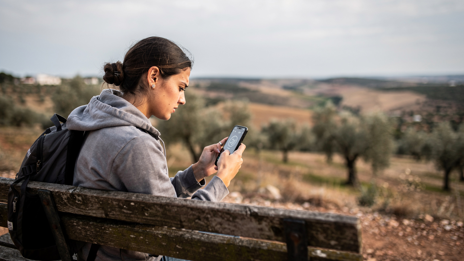 Um viajante com o semblante preocupado, sentado em um banco de madeira rústico em uma paisagem montanhosa e rural do Alentejo, Portugal, olhando para seu celular que mostra um sinal de rede fraco ou inexistente.
