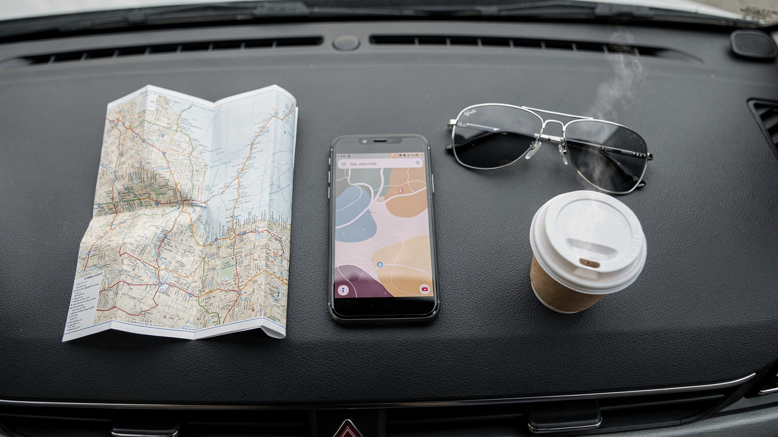 An overhead flat-lay shot on a car dashboard featuring a smartphone displaying a map, a traditional paper map folded, a pair of aviator sunglasses, and a hot coffee cup.