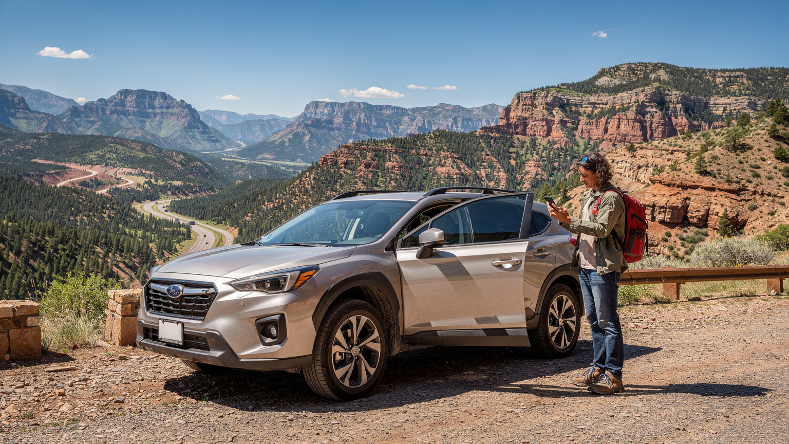 A person standing casually by the open door of their car at a scenic overlook, checking their smartphone for directions, with a breathtaking view of mountains and a winding road in the background.
