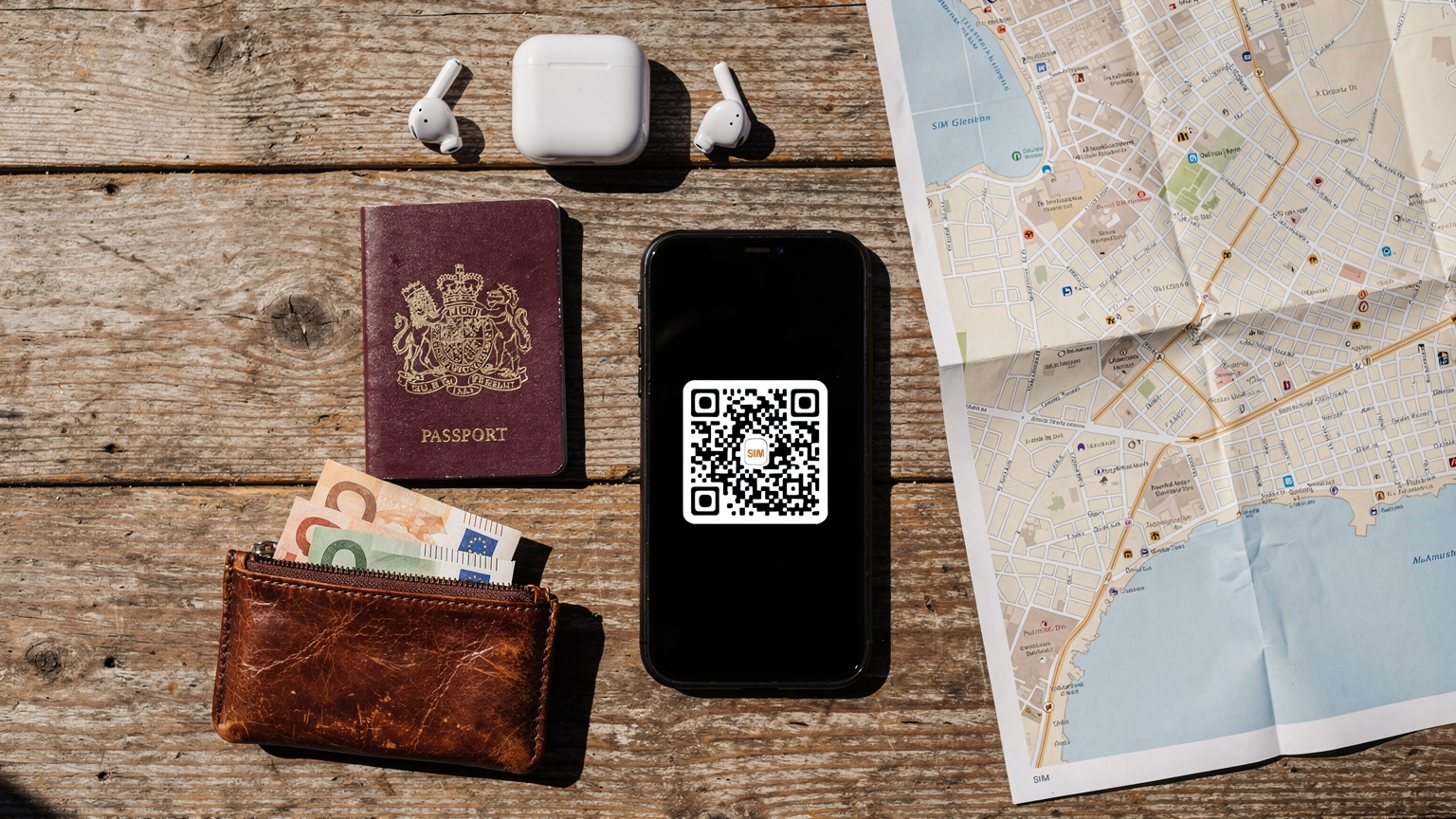 An overhead flat-lay of a traveler's essentials: a passport, a smartphone with an eSIM QR code clearly displayed on its screen, a pair of wireless earbuds, a small currency pouch with euros, and a partially unfolded map of a Mediterranean city, all neatly arranged on a rustic wooden table.