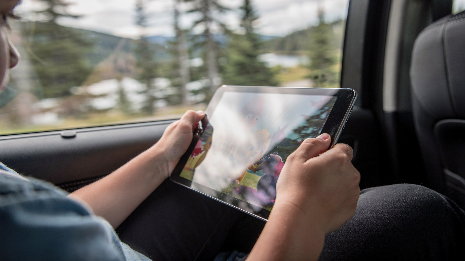 Twee kinderhanden houden een tablet vast waarop een animatiefilm wordt afgespeeld, zichtbaar in de achterbank van een auto, met een wazig landschap dat langs het raam flitst.