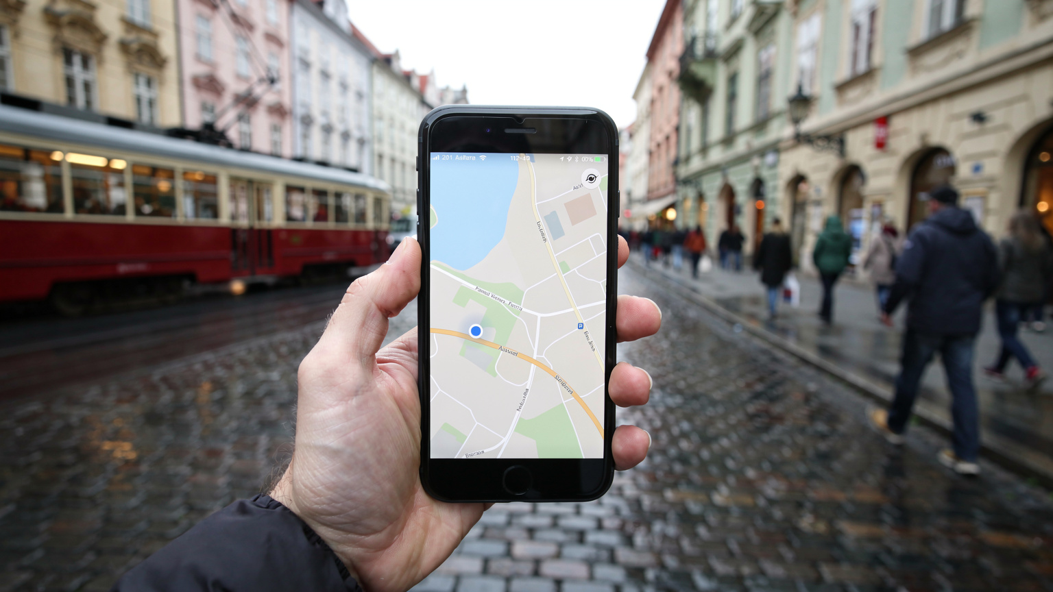 En reisende holder opp en smarttelefon med et kart over Praha på skjermen, mens en historisk trikk passerer i bakgrunnen på en brosteinbelagt gate.