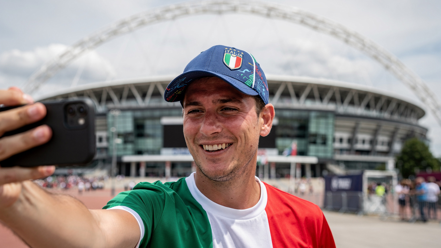 Un tifoso italiano sorride mentre si scatta un selfie davanti al famoso stadio di Wembley a Londra, tenendo in mano il suo telefono con un cappellino dell'Italia in testa, simbolo di gioia e connessione.