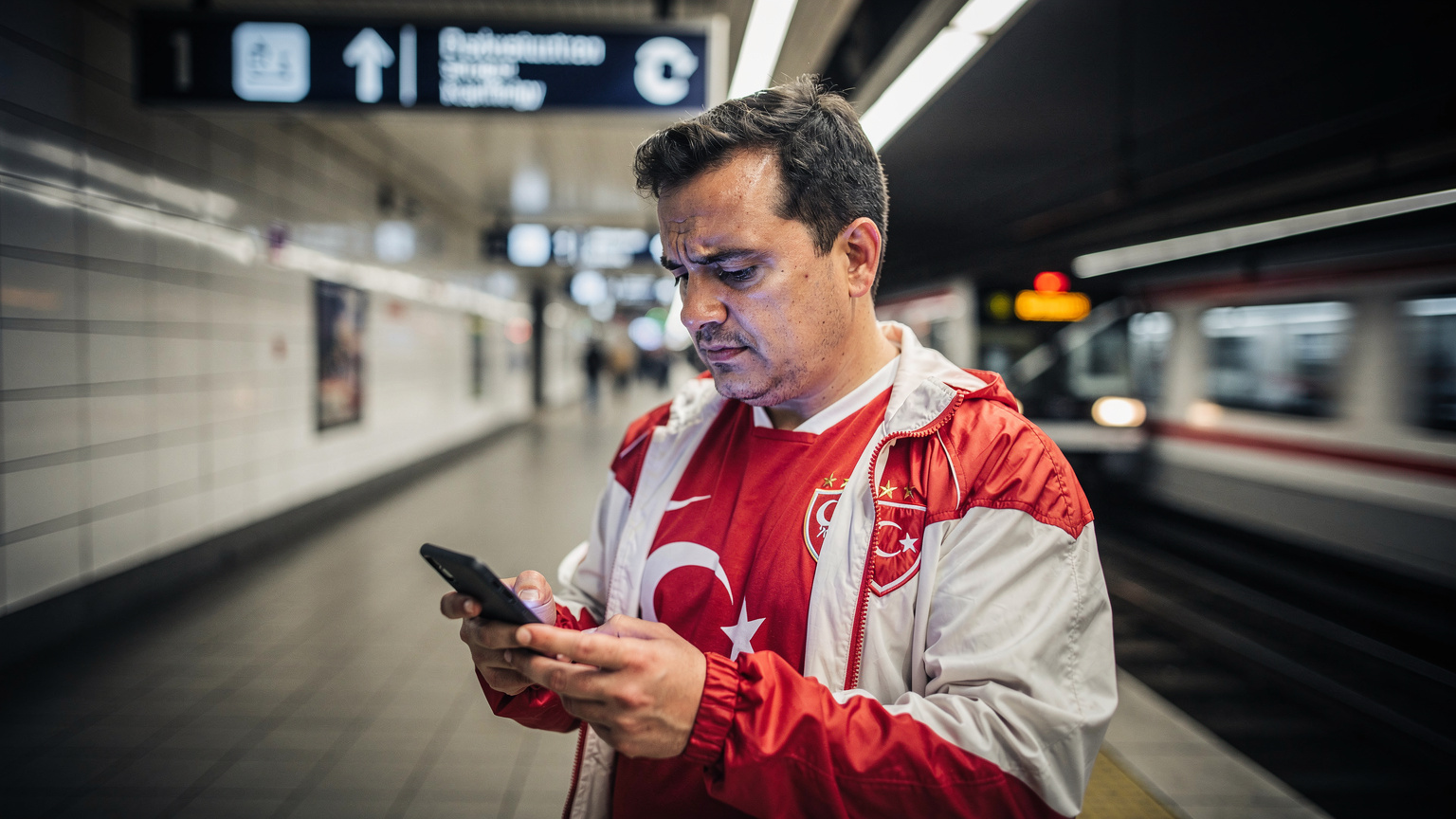 Almanya'da bir U-Bahn istasyonunda, akıllı telefonunda bir harita uygulamasını dikkatle inceleyen ve hafifçe kaşları çatılmış bir Türk taraftar.