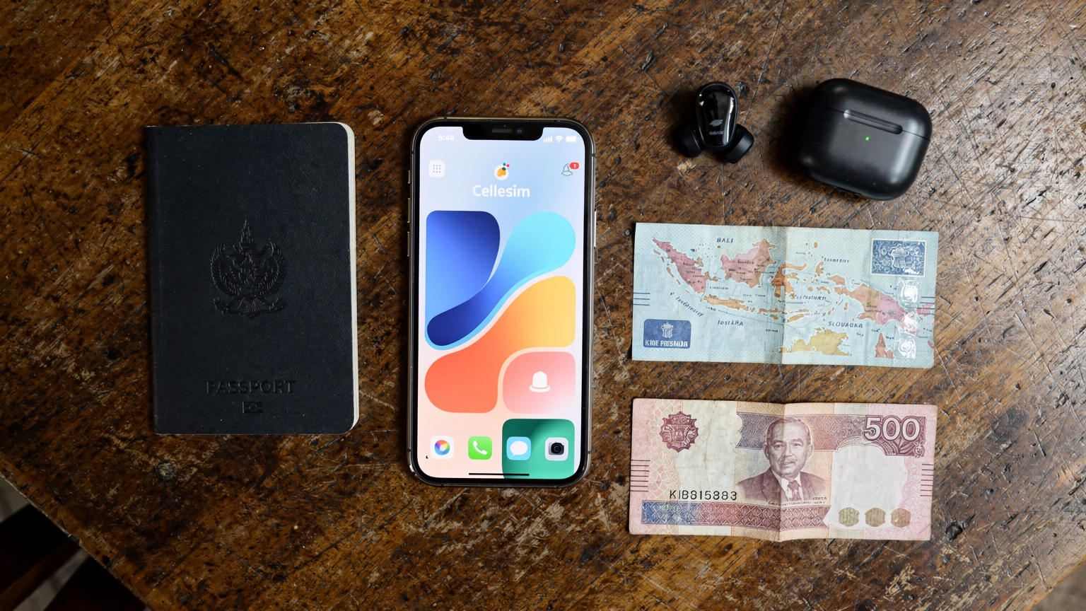 An editorial overhead flat-lay of travel essentials: an iPhone 17 with an active Cellesim eSIM, a passport, wireless headphones, a small map of Bali, and a local currency note, all on a weathered wooden table.