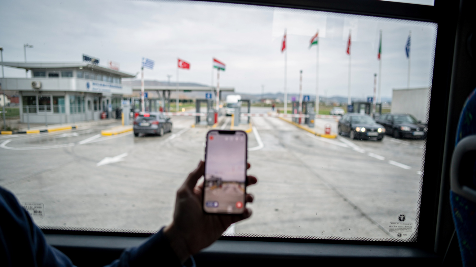 Bir otobüs penceresinden görünen, Türkiye-Yunanistan veya Türkiye-Bulgaristan sınır kapılarından birinin manzarası. Otobüs sınır kontrolü için yavaşça ilerliyor. Pencerenin kenarında, elinde akıllı telefon tutan bir yolcunun bulanık silüeti var, telefon ekranı hafifçe parlıyor.