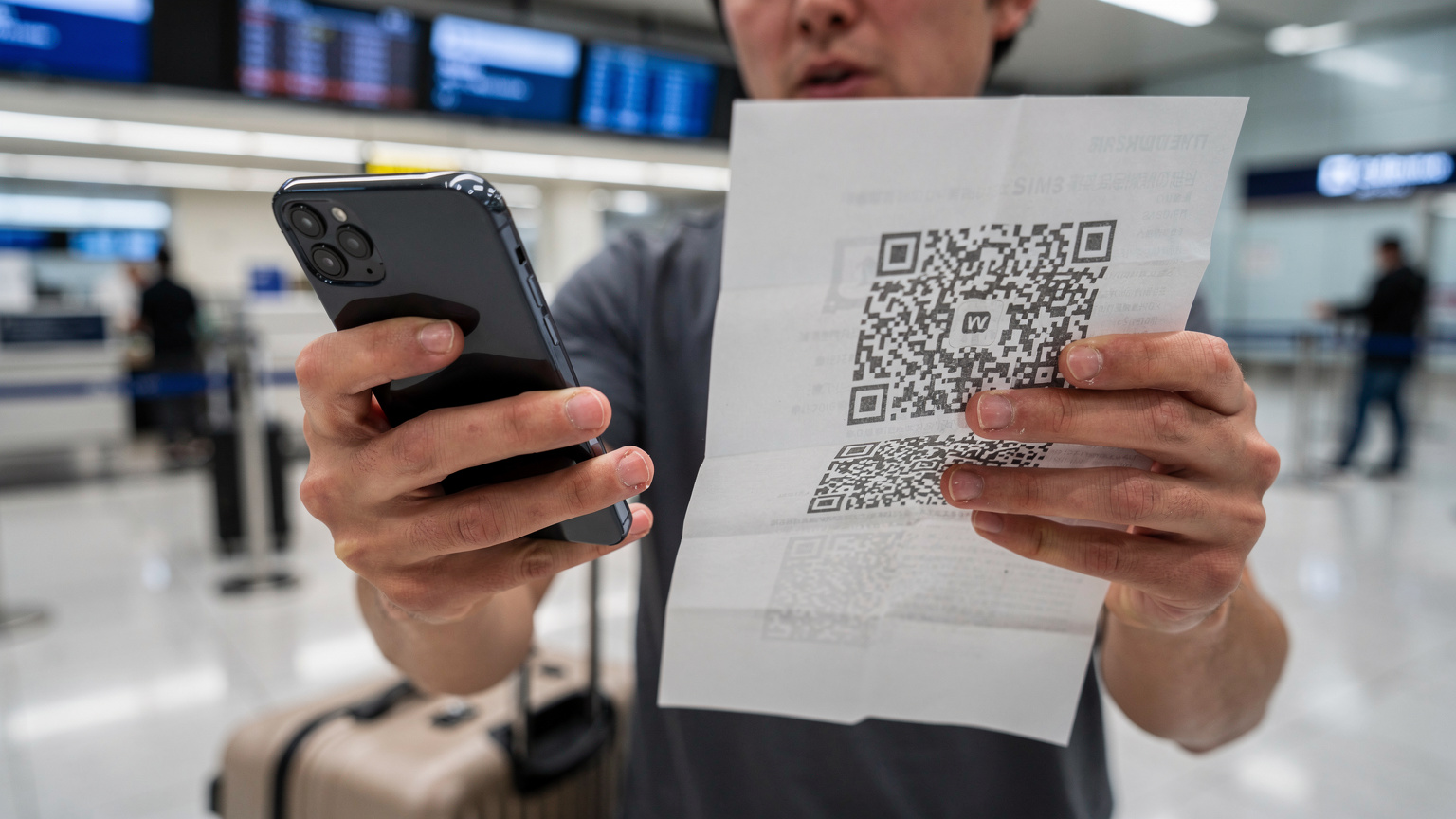 空港のチェックインカウンターで、スマートフォンでeSIMのQRコードを読み込もうとしている日本人旅行者の手元。焦りの表情が見える。