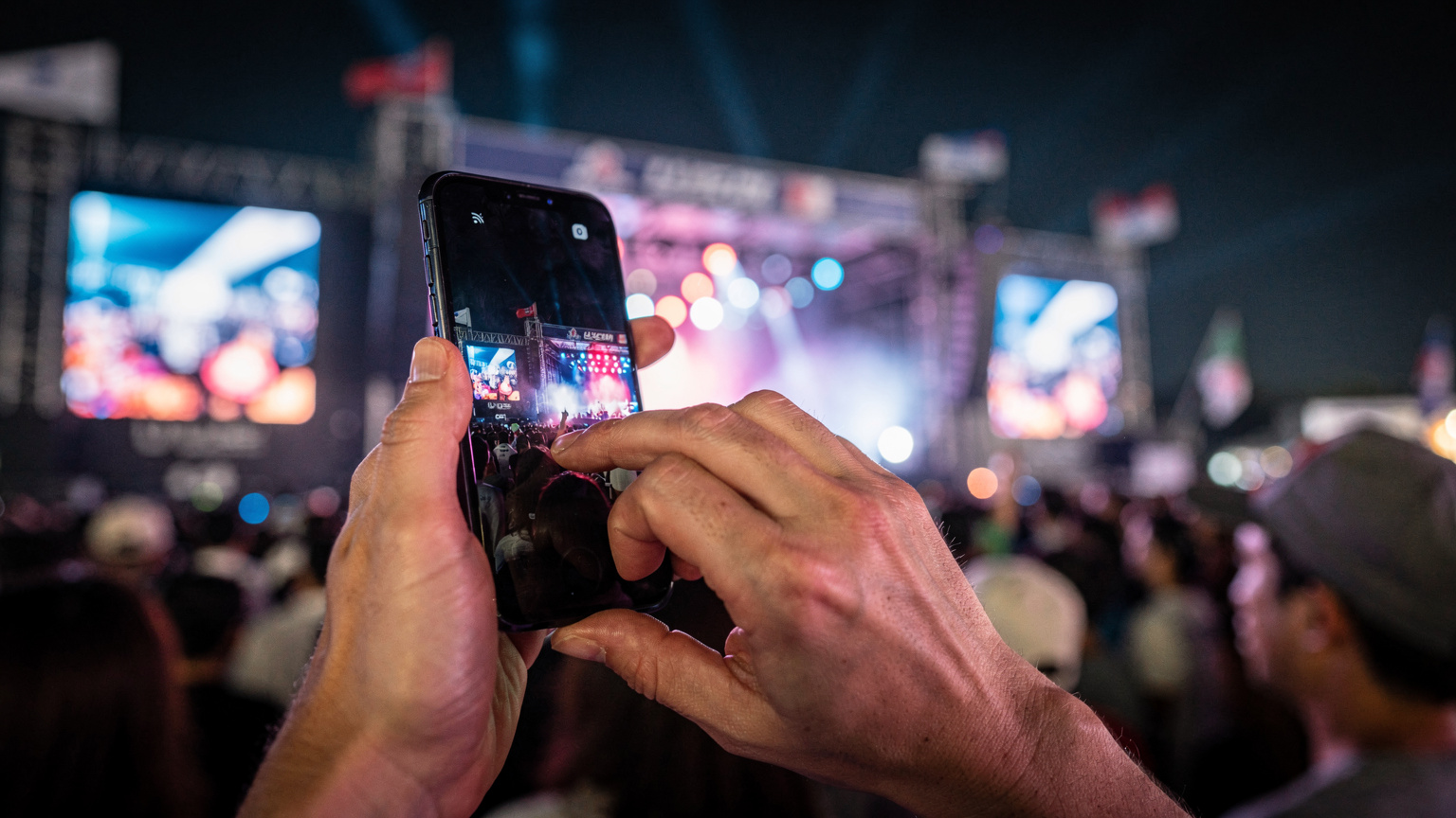 韓国の活気ある音楽フェスで、スマートフォンを操作する日本人旅行者の手元。ステージの光が背後に滲む。