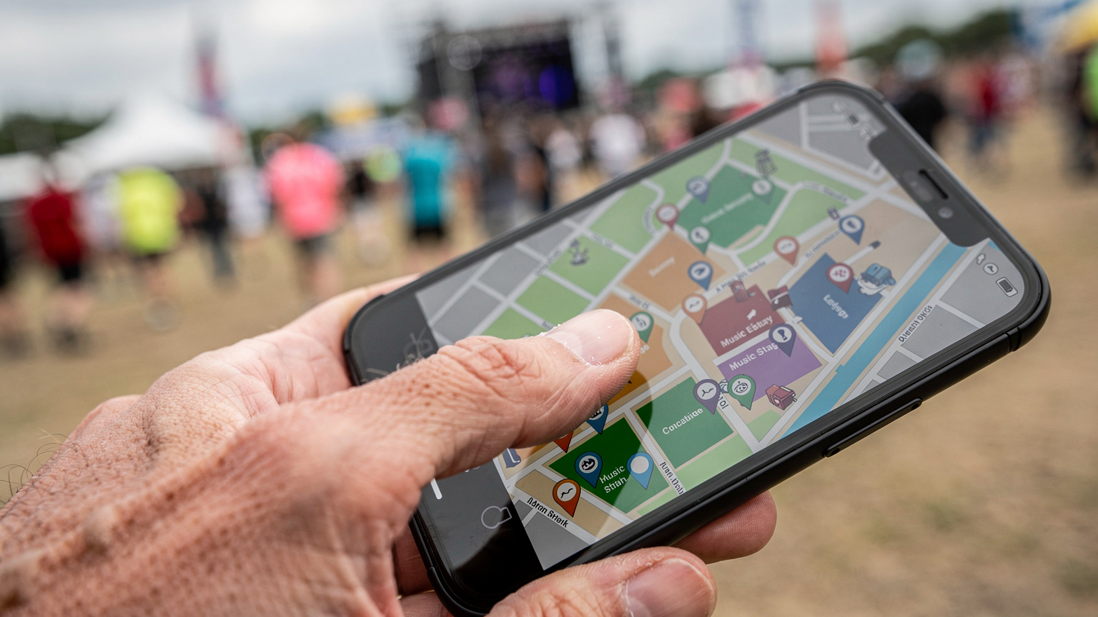 Ręka dorosłej osoby, delikatnie dotykająca ekranu smartfona, na którym widać mapę festiwalu z zaznaczonymi scenami i punktami orientacyjnymi. W tle rozmyte postacie idące w tłumie.