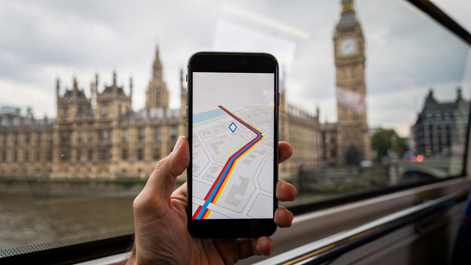 Una mano sostiene un teléfono móvil mostrando una aplicación de mapa con una ruta en un autobús turístico de dos pisos en Londres, con el Big Ben al fondo, bajo un cielo nublado.