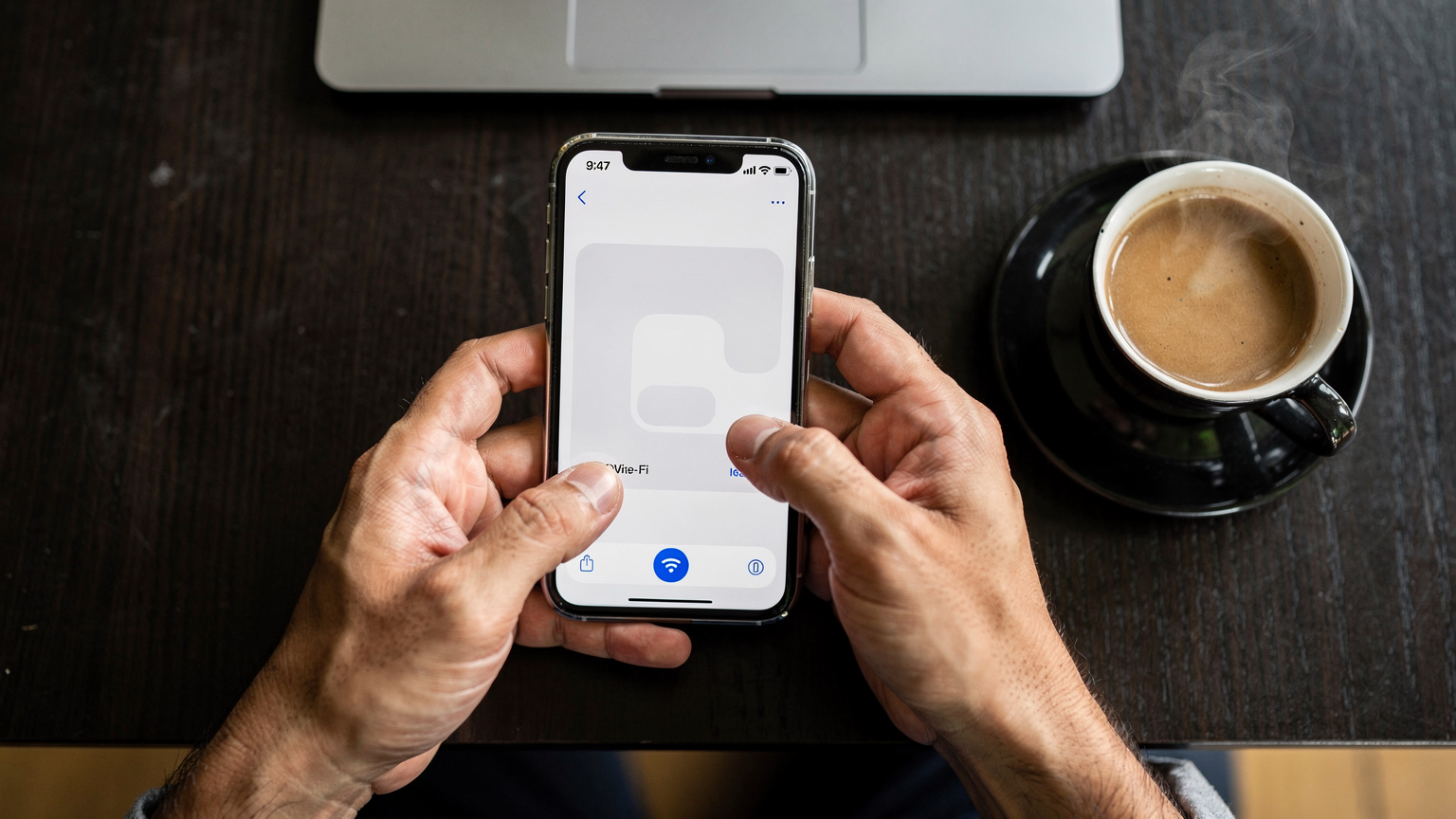 Tangan seorang pria sedang memegang iPhone dengan layar pengaturan Wi-Fi terbuka, di meja kayu gelap dengan latar belakang laptop dan secangkir kopi, di sebuah co-working space di Bandung.
