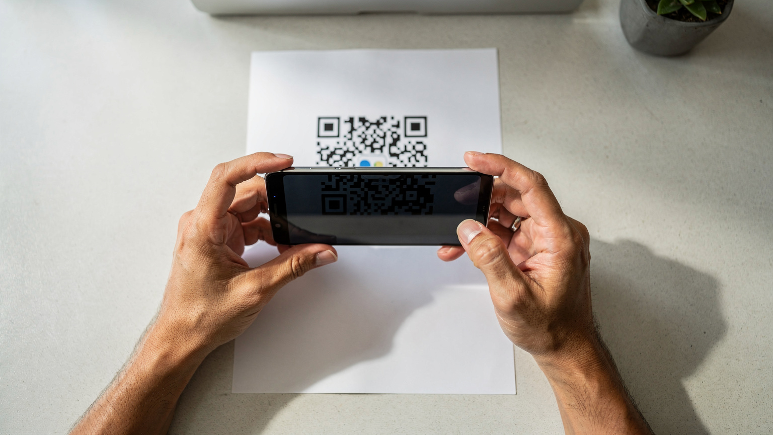 Tangan seseorang memegang smartphone modern yang sedang memindai kode QR eSIM yang tercetak di selembar kertas.