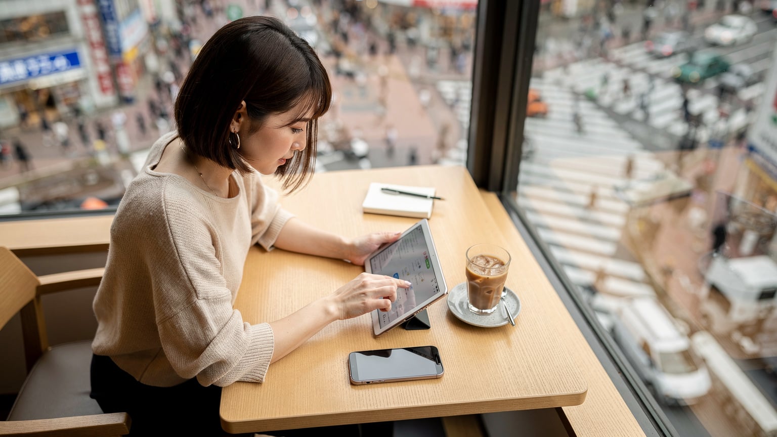 渋谷のスクランブル交差点を見下ろすカフェで、タブレットとスマートフォンを並べて旅行計画を練る日本人女性。eSIMプランの比較画面を開いている。