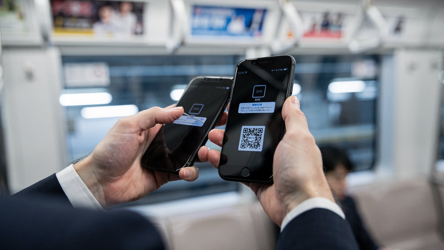 東京の地下鉄車内で、スマートフォンでeSIMのQRコードをスキャンして設定する日本人ビジネスマンの手元。背景には窓外の景色がぼんやりと映る。