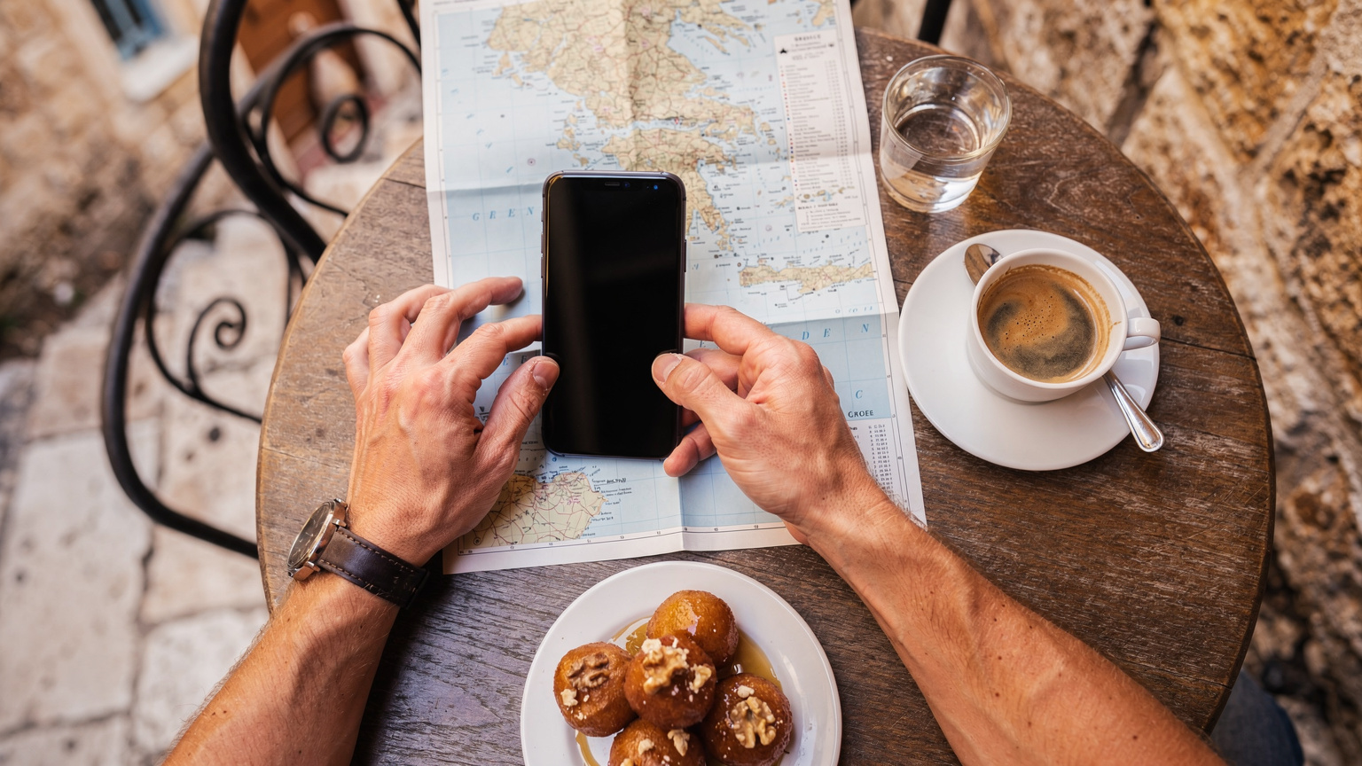 En tæt på-optagelse af en rejsende, der holder sin smartphone op mod et kort over Grækenland og navigerer, mens hun sidder på en caféterrasse i Chania, Kreta, med en kop græsk kaffe ved siden af.