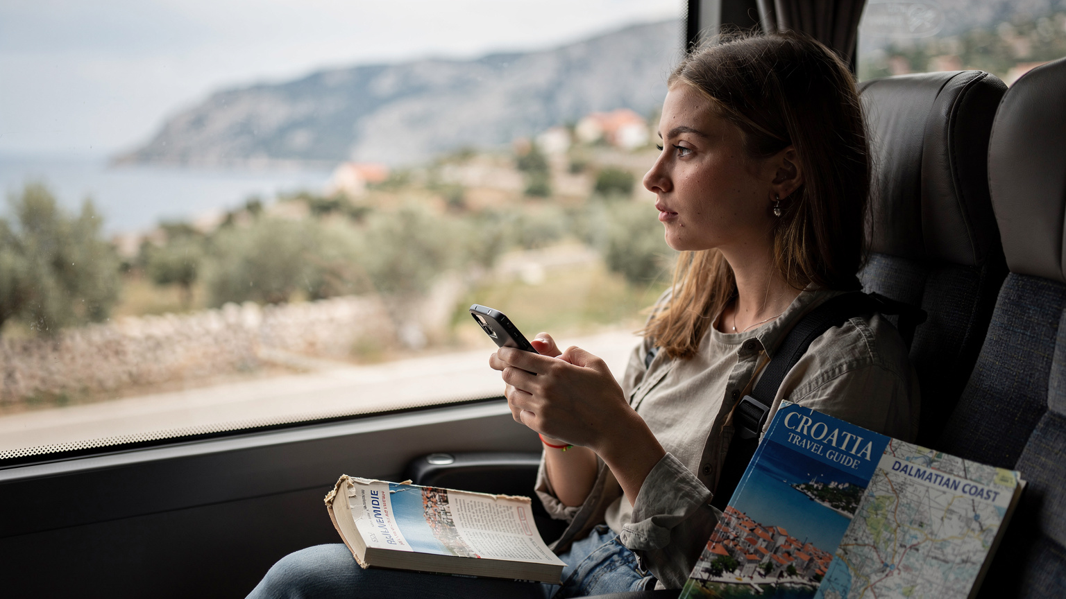 Cestovatelka se s mobilem v ruce dívá z okna autobusu na chorvatskou krajinu. Před sebou má průvodce a mapu, kontroluje si spojení.