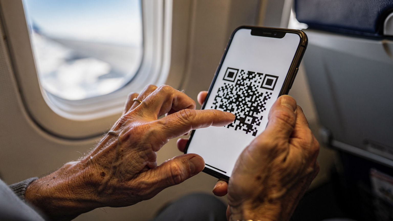 A close-up shot of an older traveler's hands gently tapping on a smartphone screen, which displays an eSIM activation QR code, with a blurred airplane window and wing visible in the background.