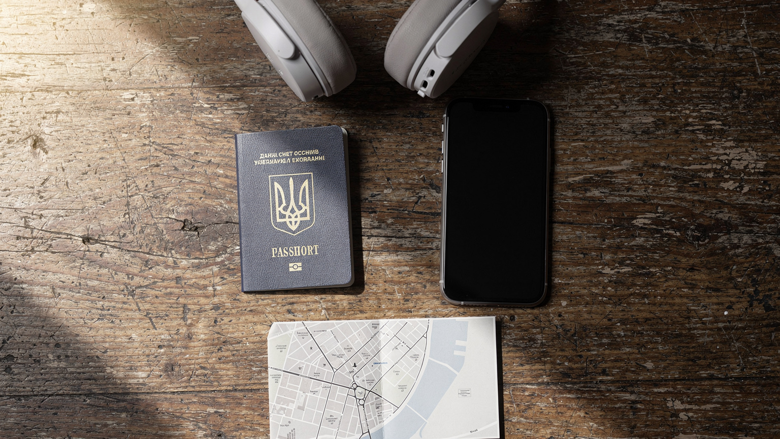 Плоске зображення на дерев'яному столі: український паспорт, смартфон, навушники та невеликий шматочок карти, що символізують підготовку до подорожі.