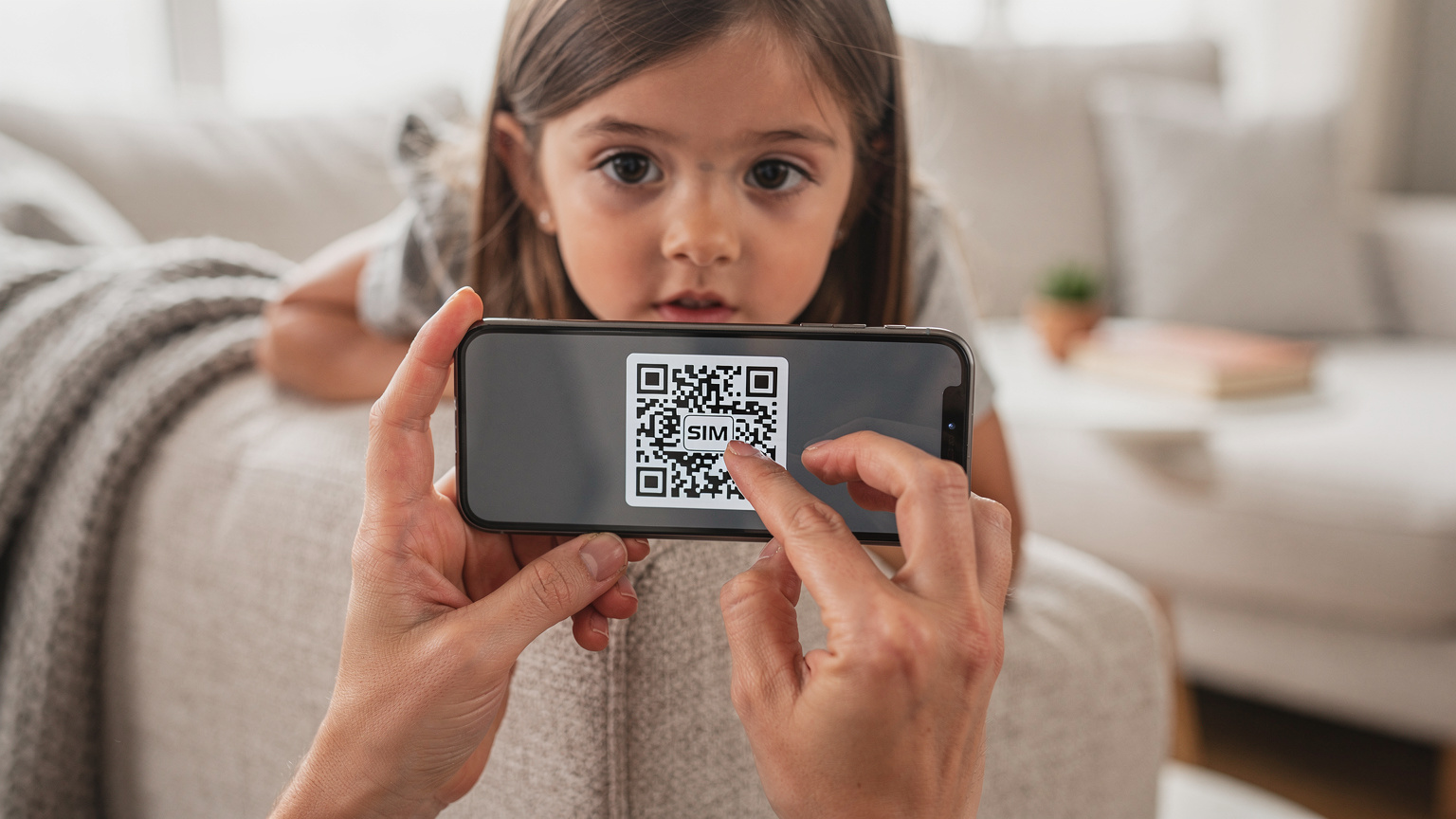 Tangan seorang ibu menunjuk ke arah telefonnya yang memaparkan kod QR eSIM untuk dipasang, sambil anak perempuannya yang lebih muda melihat dengan rasa ingin tahu. Latar belakang menunjukkan ruang tamu rumah yang kemas.