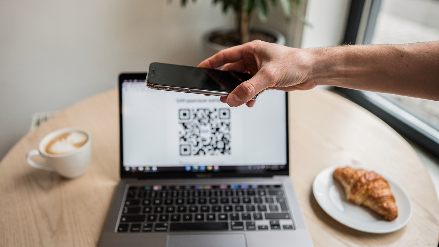 O mână de om scanează un cod QR de pe un laptop deschis, care afișează instrucțiuni de activare eSIM, într-o cafenea modernă și luminoasă din Berlin.