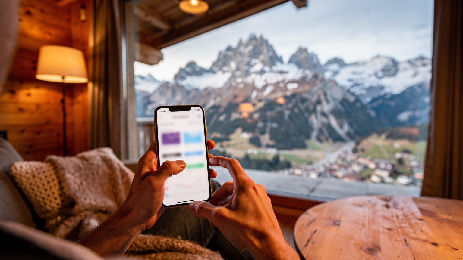 Un călător folosește o aplicație mobilă pentru a-și monitoriza consumul de date eSIM pe un telefon, având în fundal o fereastră care arată o vedere panoramică asupra Alpilor Elvețieni de la o cabană montană.