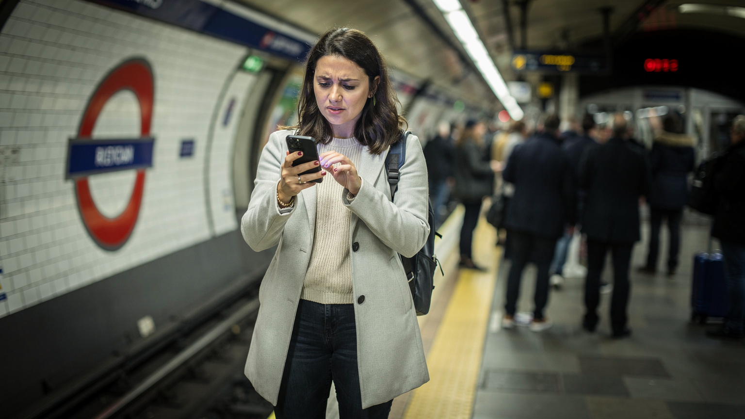 Una donna italiana con un'espressione frustrata guarda lo schermo del suo smartphone in una stazione della metropolitana di Londra affollata, con i volti sfocati di altri pendolari sullo sfondo.
